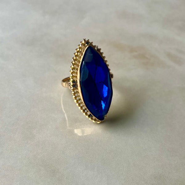 A blue stone cocktail ring with a gold band, viewed from the side on a reflective surface.