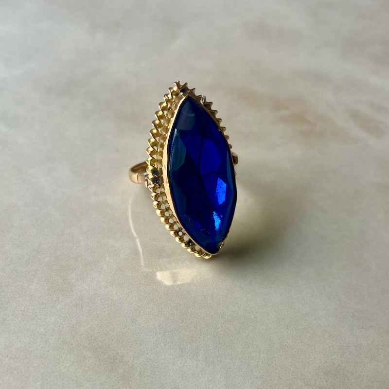 A blue stone cocktail ring with a gold band, viewed from the side on a reflective surface.