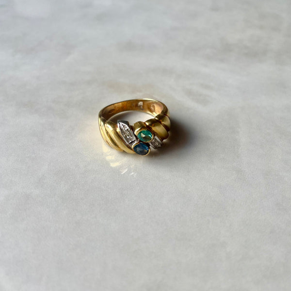 An 18 carat yellow gold ring with a white gold setting, featuring a central sapphire flanked by a diamond and emerald, set on a marble surface.
