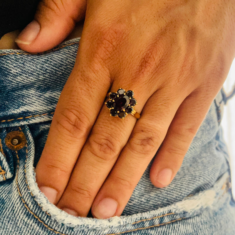 Hand wearing a decorative ring with garnet stones and gold accents, against a denim background | Deheb Jewellery Malta