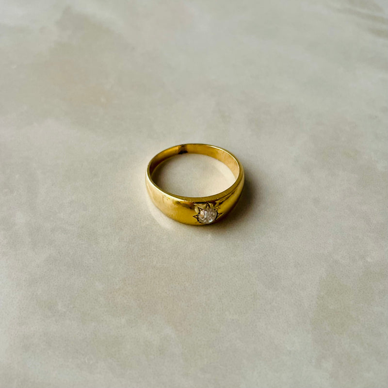 A single gold gypsy ring with a central old cut diamond, displayed on a marble surface.