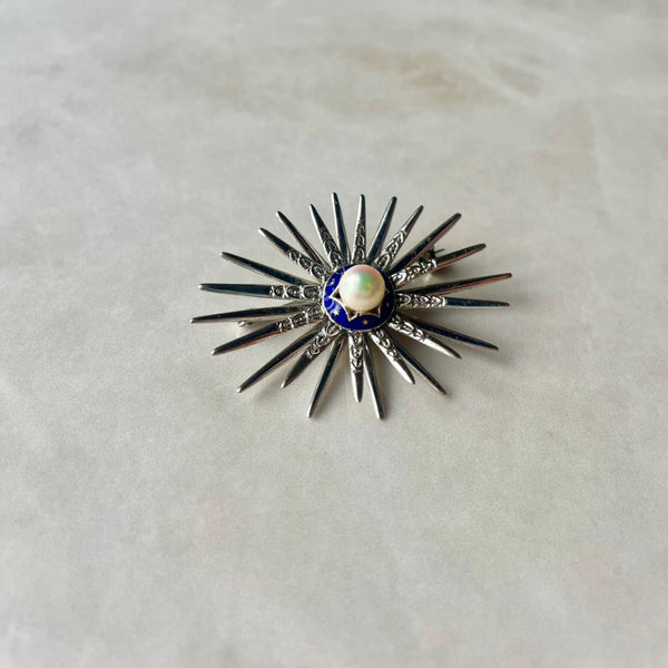 A star-shaped brooch made of 18 carat white gold, with a central cultivated pearl and blue enamel detail at the center.
