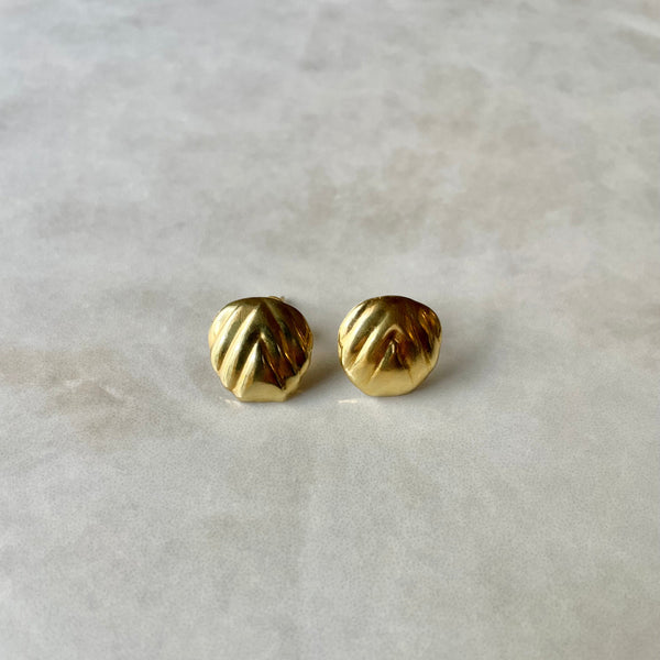A pair of round ripple, gold stud earrings with ridged detailing, displayed against a marble background.