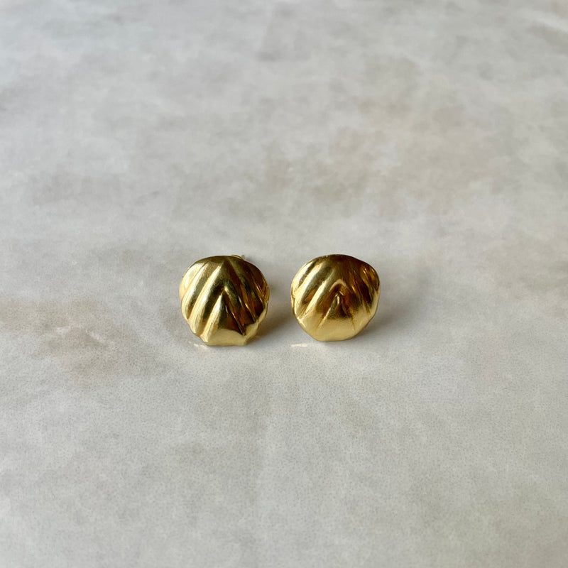 A pair of round ripple, gold stud earrings with ridged detailing, displayed against a marble background.