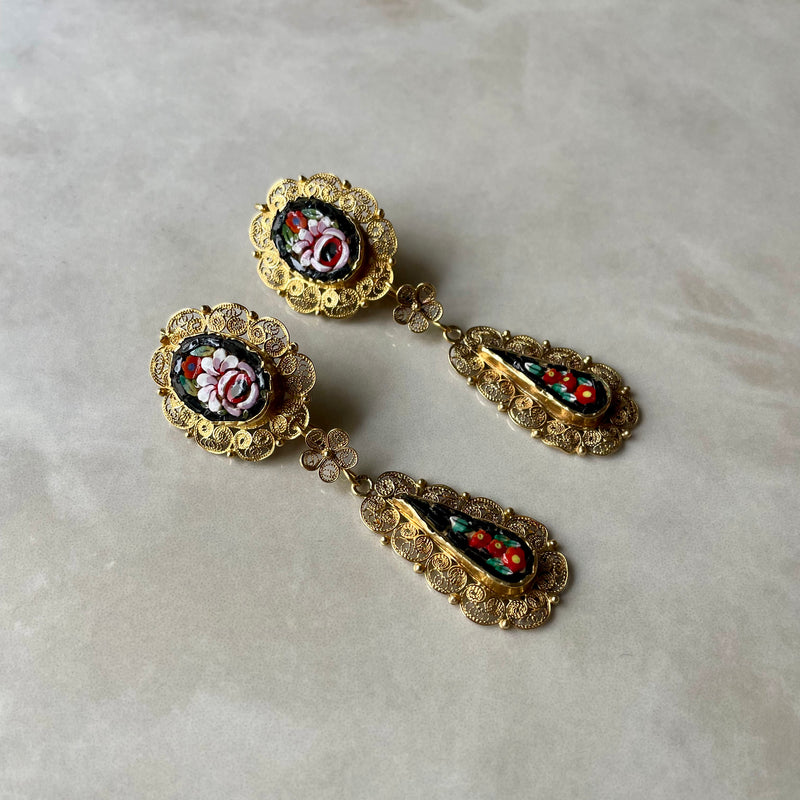 A pair of intricate gold filigree earrings with micro mosaic details, placed on a marble surface.