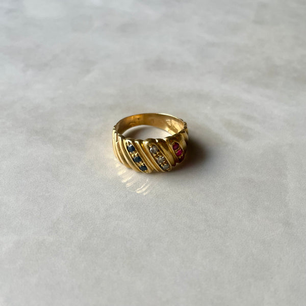 A gold ring with three rows of stones, including rubies, sapphires, and diamonds, set against a marble background.